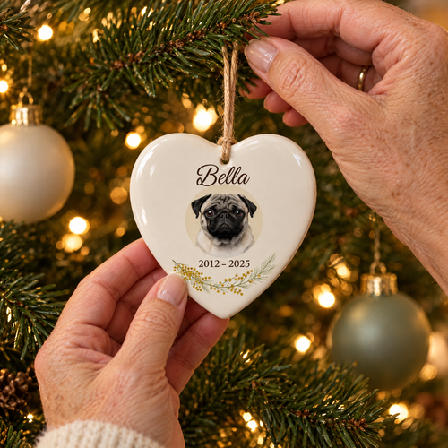Hands hanging a personalised ceramic heart pet memorial ornament with a pug named Bella, 2012 to 2025, on a lit Christmas tree